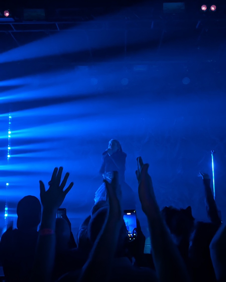 Hayla performing live, lit in blue stage light, with the crowd's hands raised in front of the stage